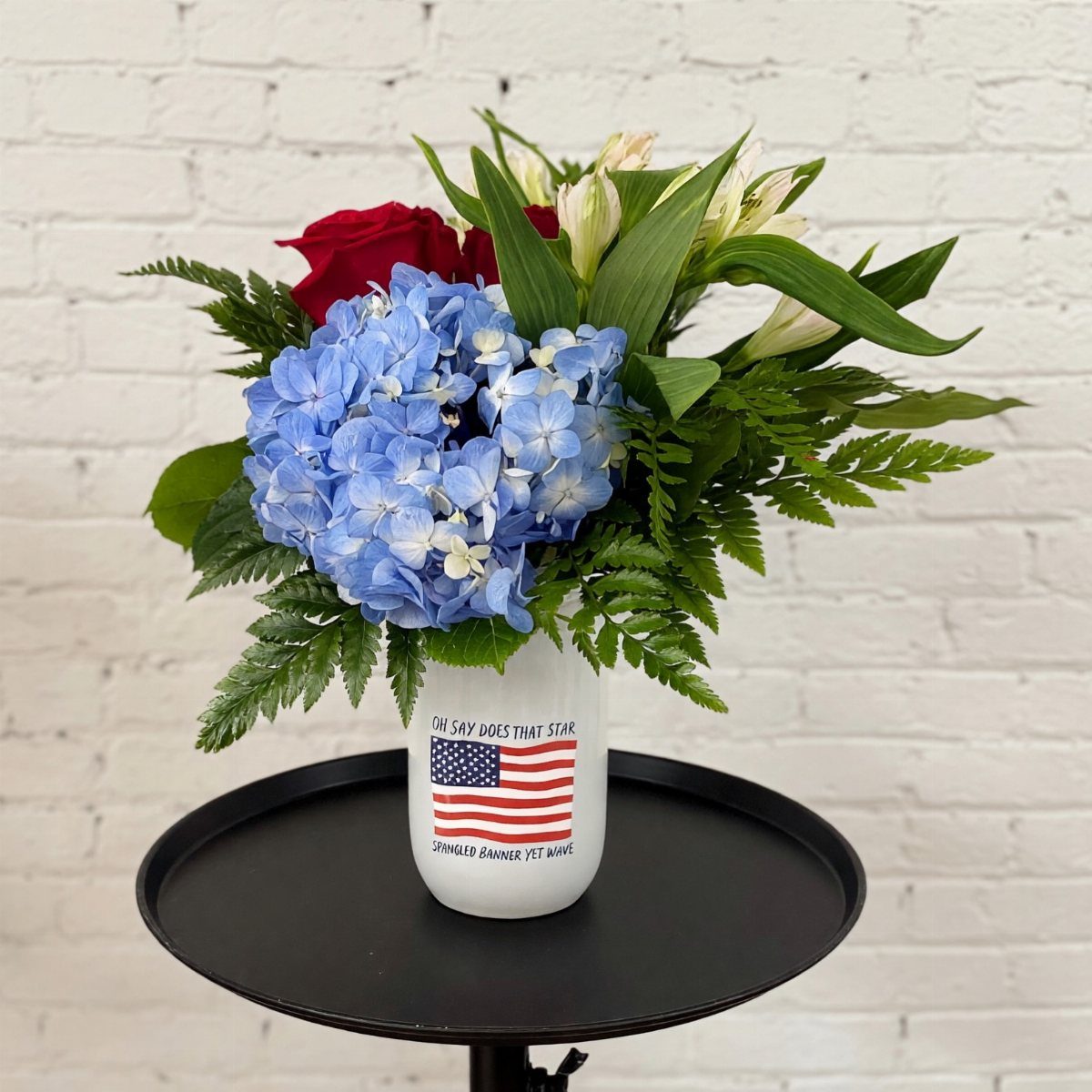 White vase with American flag holding red, white, and blue floral arrangement.