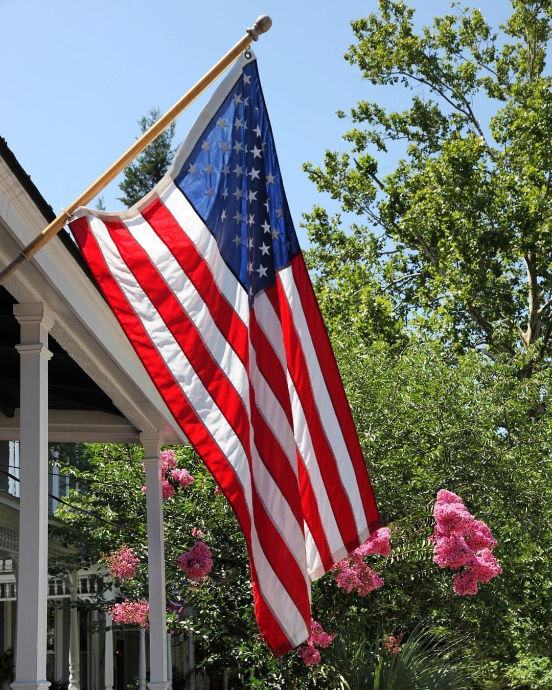 American flag hanging from a white porch with green trees and pink flowers.