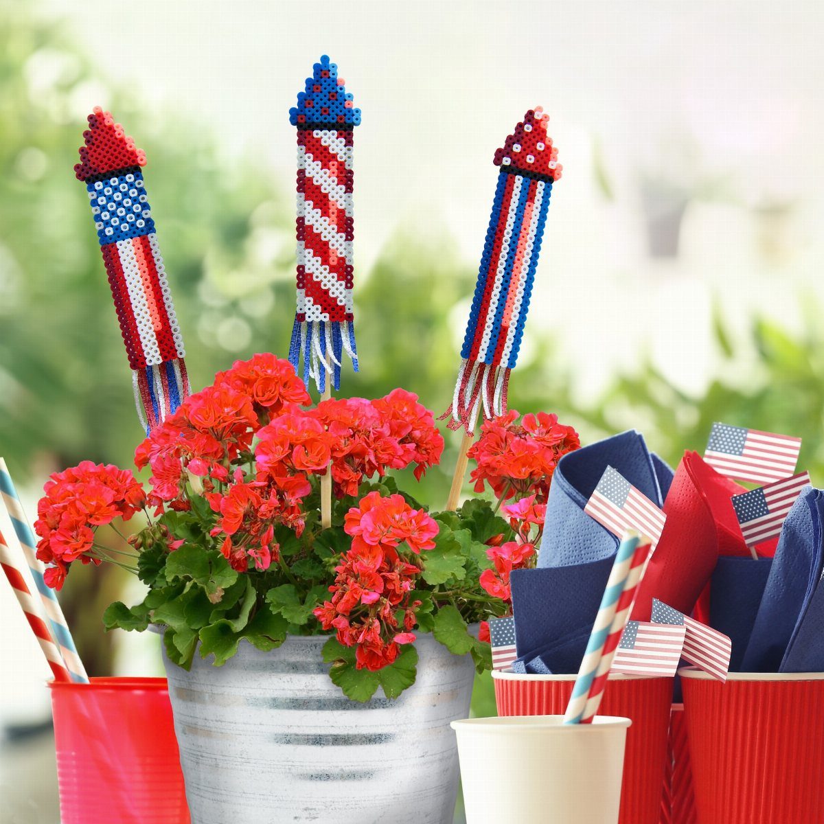 Beaded rocket stakes in a metal bucket of red flowers with patriotic accessories.