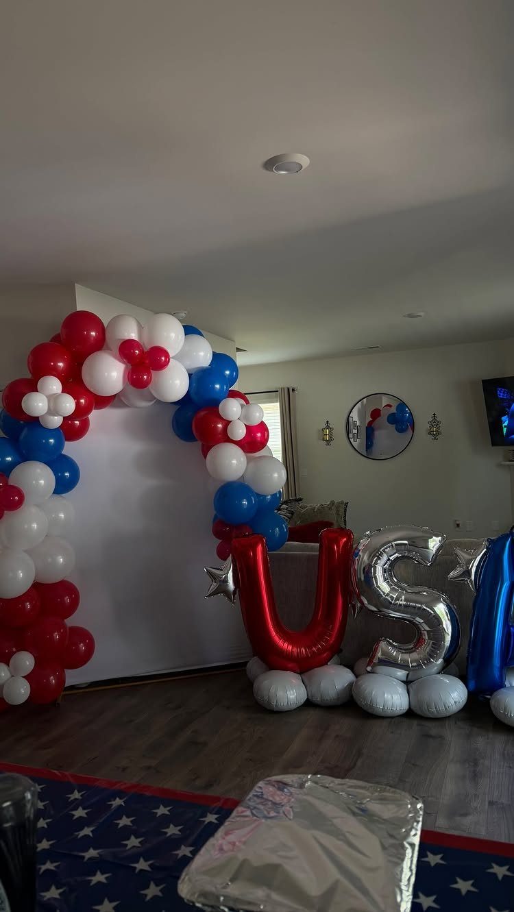 Red, white, and blue balloon arch with large USA foil balloons indoors.