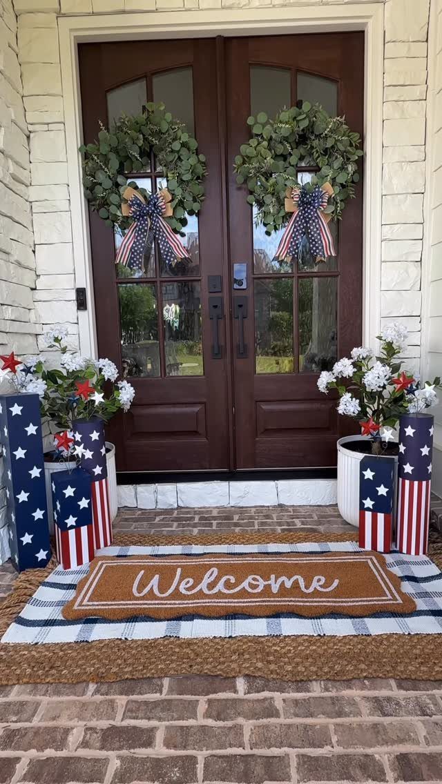 Patriotic porch decor with flag wreaths, red white and blue flowers, and welcome mat.