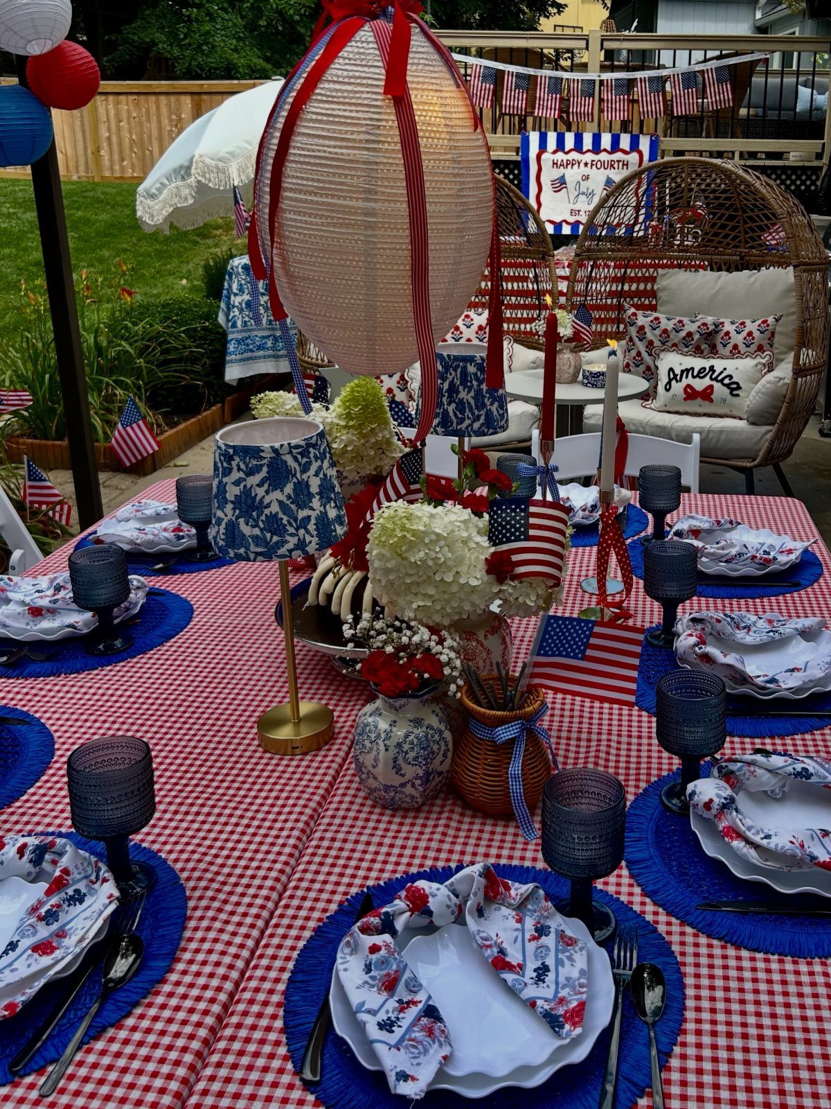 Patriotic table setting with red checkered cloth, blue accents, and American flags outdoors.