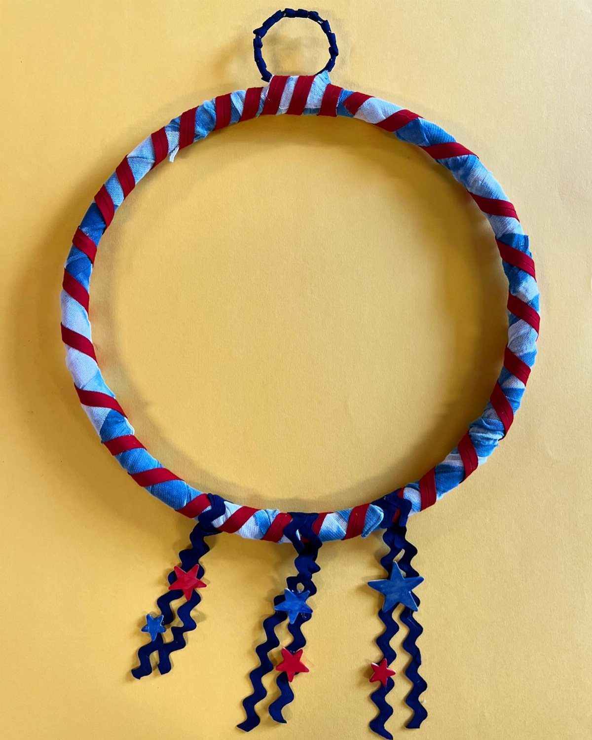 Red, white, and blue fabric wrapped wreath with hanging star ribbons on yellow wall