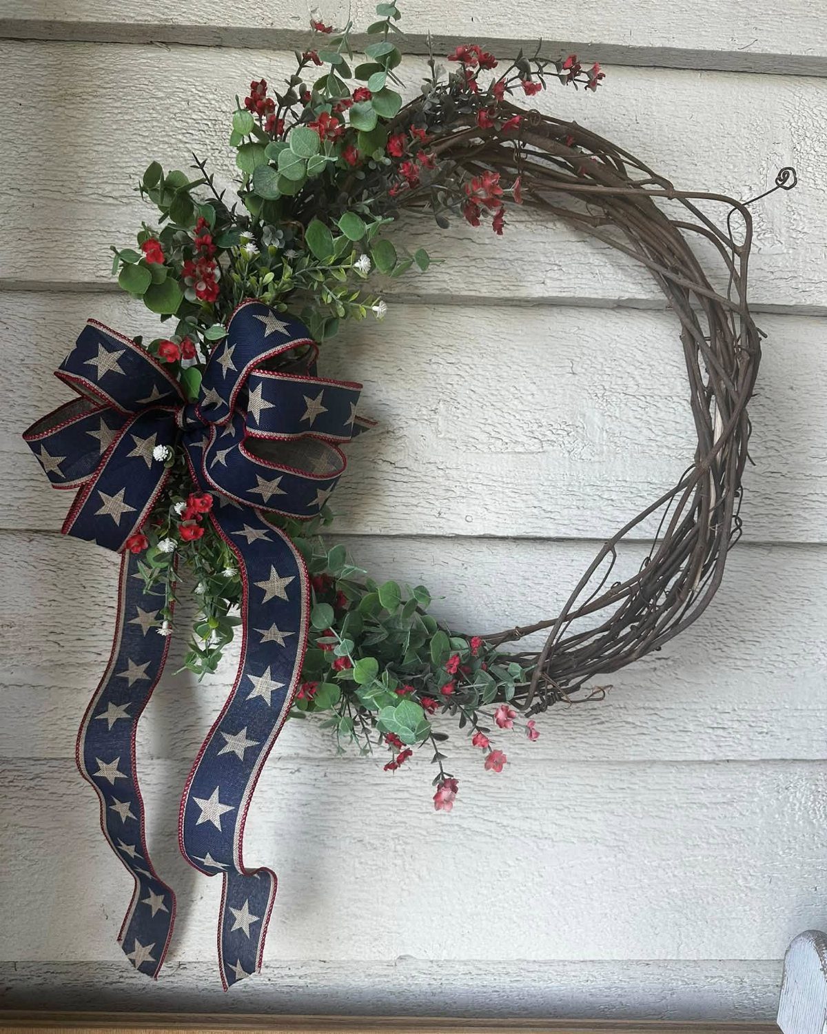 Grapevine wreath with green leaves, red berries, and a star-patterned patriotic ribbon.