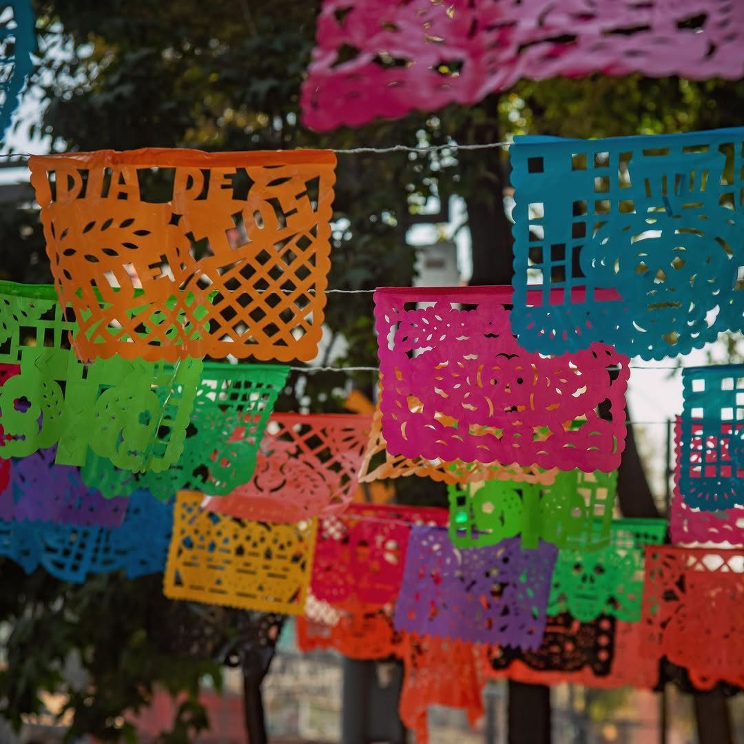 Colorful papel picado banners with intricate cutouts hanging outdoors against green trees.