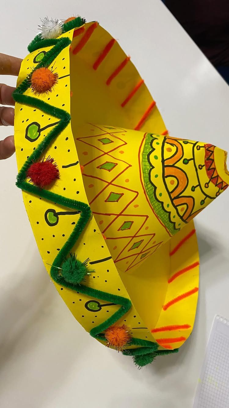 Handmade yellow paper sombrero with colorful pom-poms and green zig-zag trim decorations.