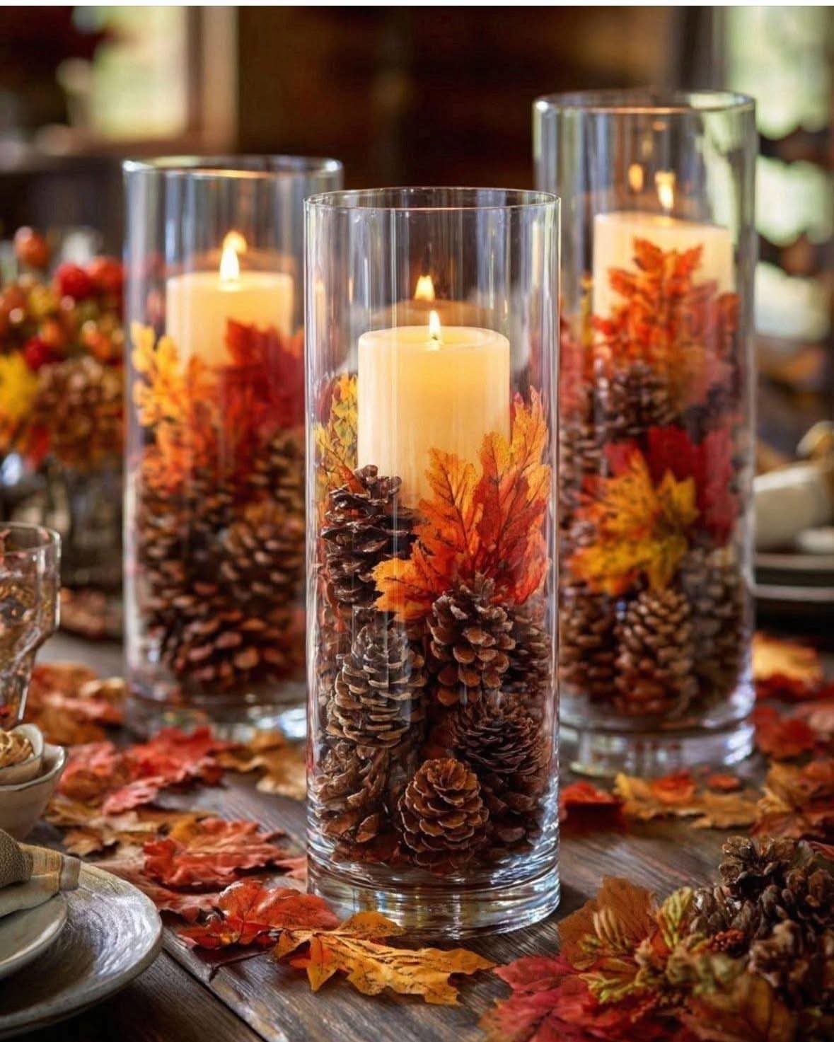 Three glass cylinder vases with white candles, pinecones, and autumn leaves on wooden table