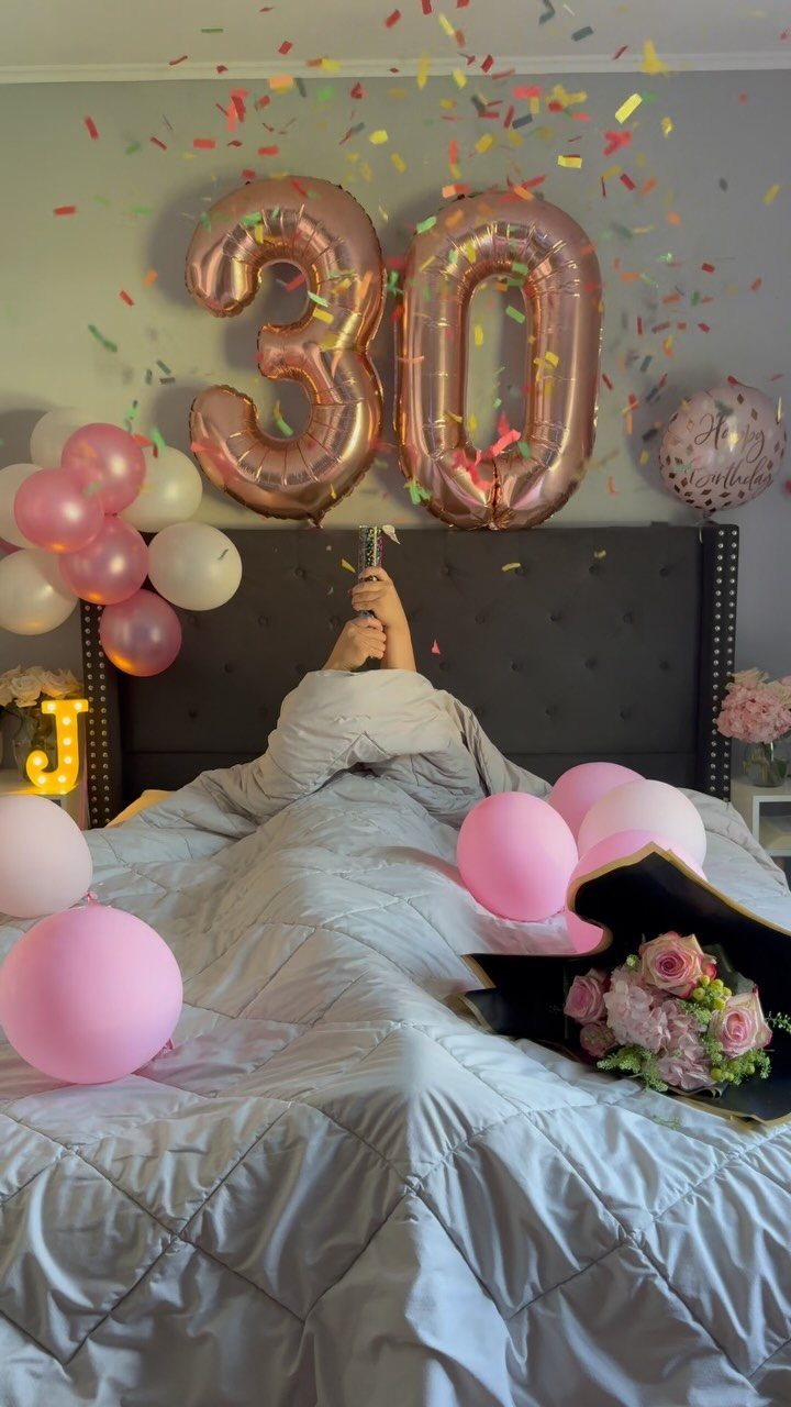 Person in bed surrounded by pink balloons and rose gold 30 balloons with confetti.