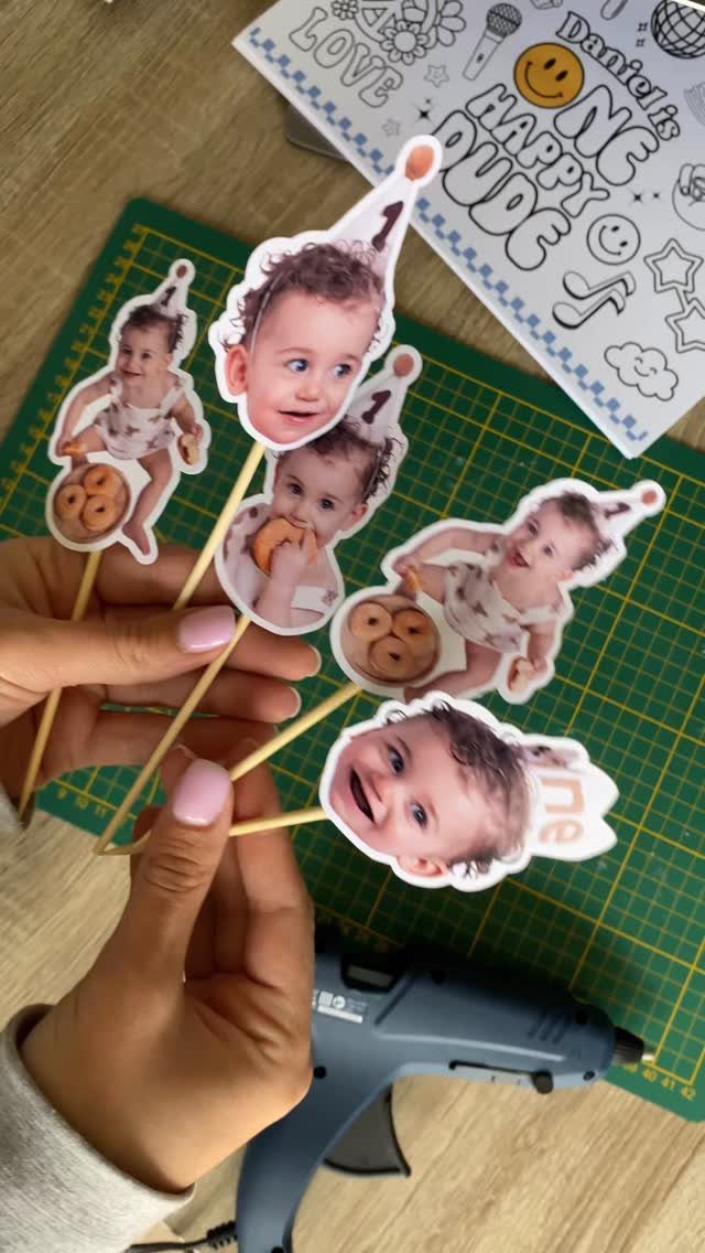 Custom cake toppers with baby photos held over a green crafting mat.