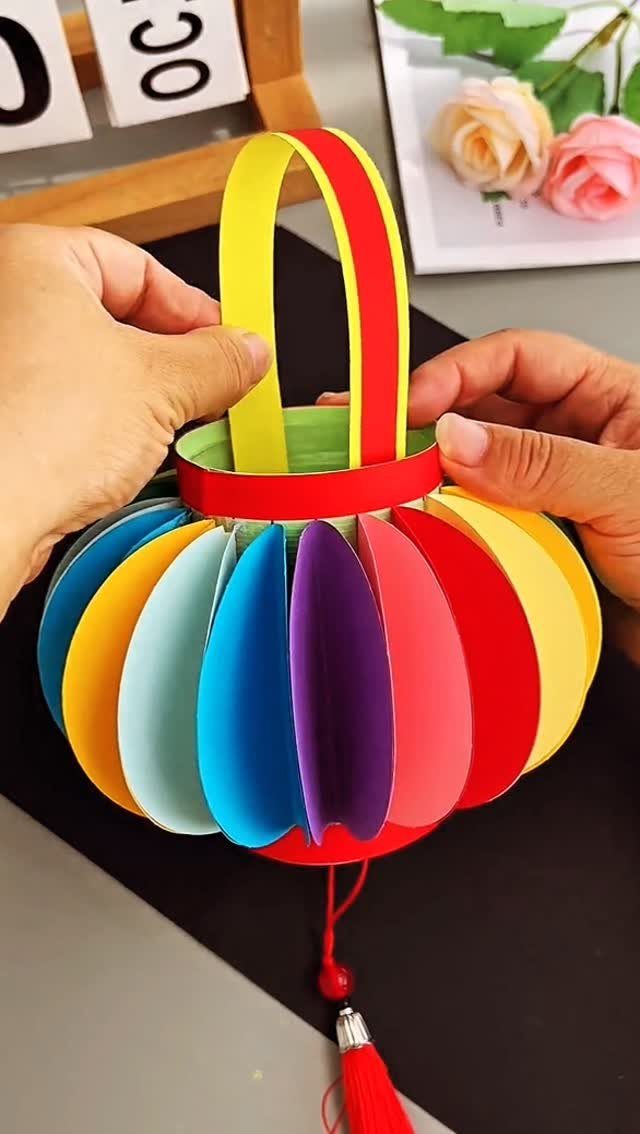 Colorful paper lantern with a yellow handle and red tassel held by hands.