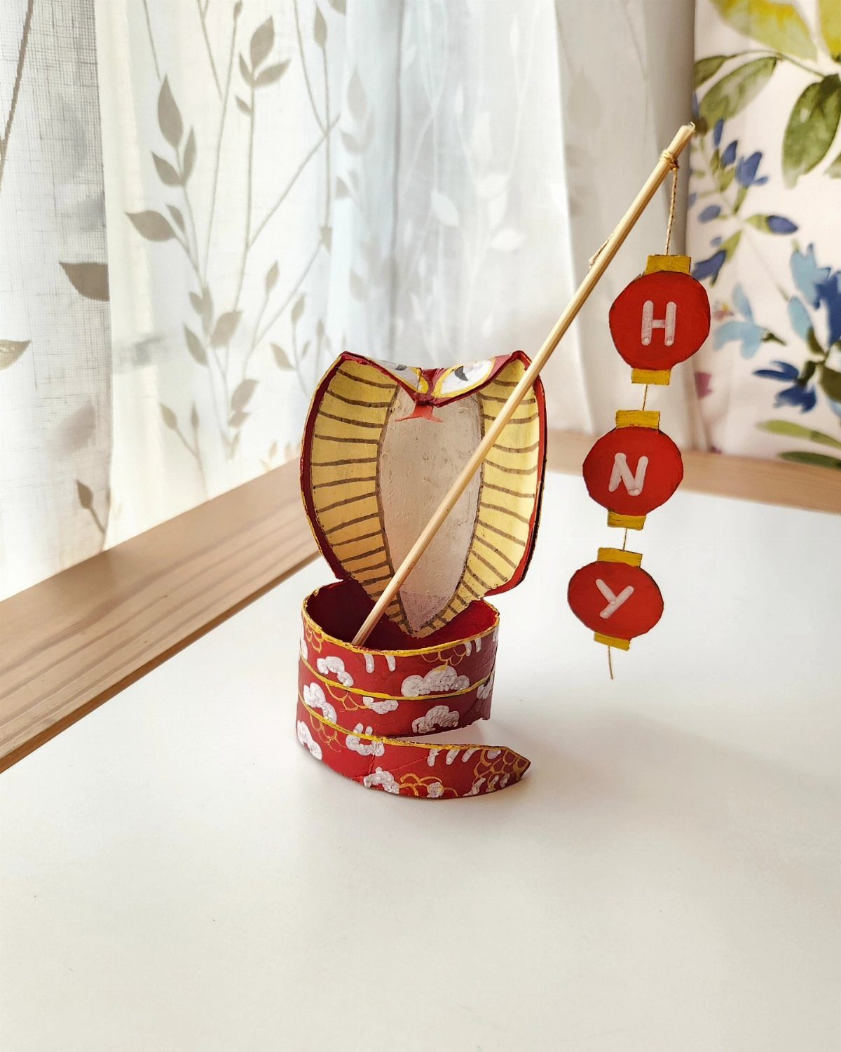 Handmade cardboard tube snake craft with red lanterns spelling HNY on a stick.