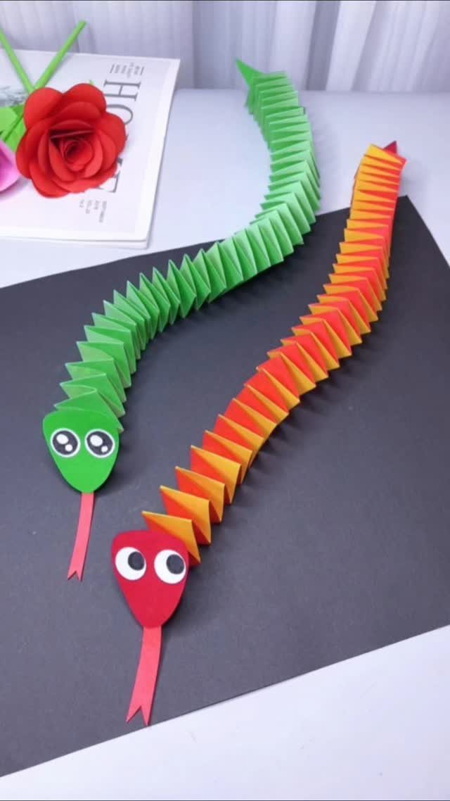 Two colorful paper snakes with googly eyes on a dark grey background.