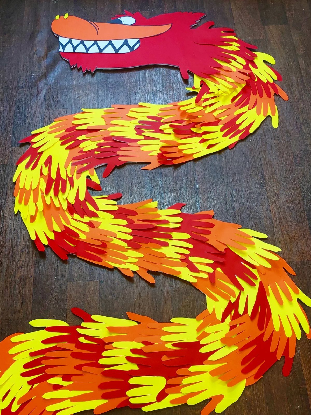 Colorful paper handprint dragon with red, orange, and yellow scales on wood floor