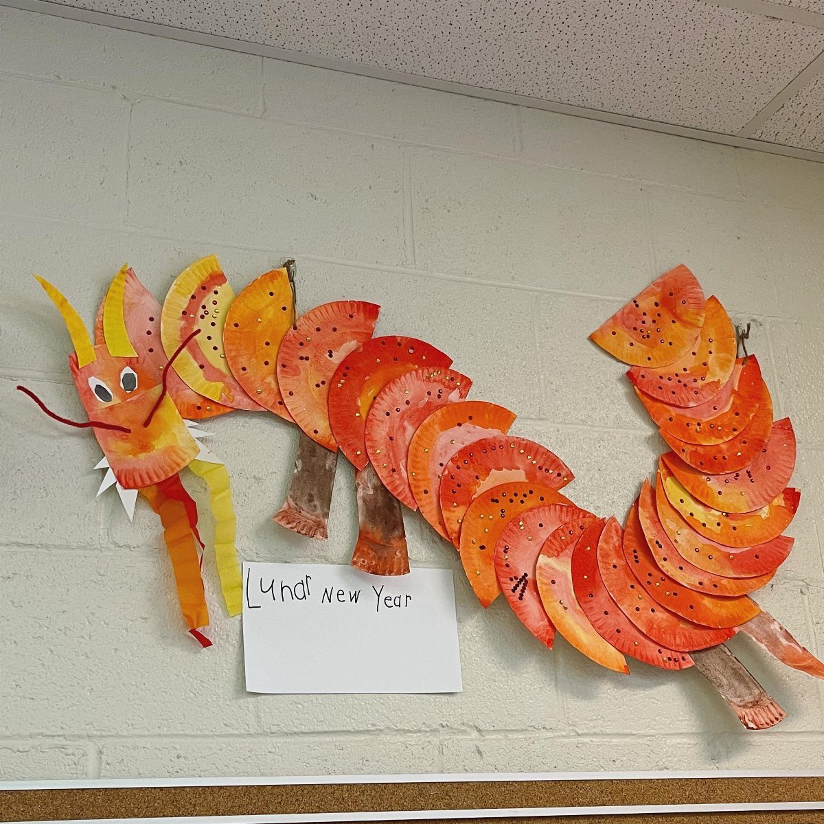 Orange and yellow paper plate dragon craft displayed on a white brick wall.
