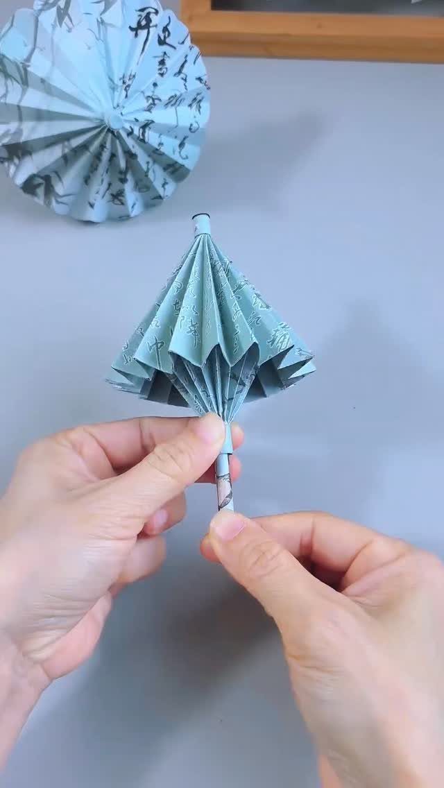 Hands assembling a small light blue pleated paper umbrella with calligraphy patterns