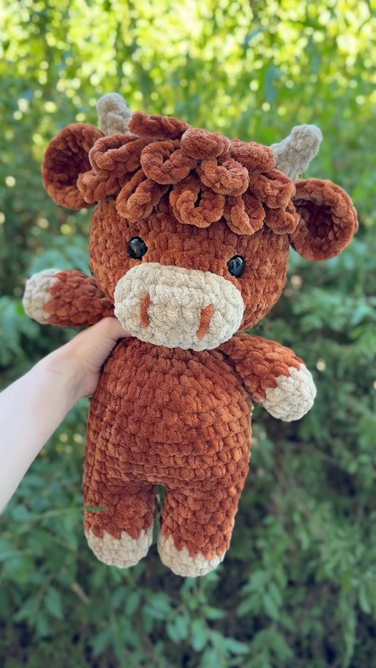 Brown crochet plush cow held by a hand against a green leafy background