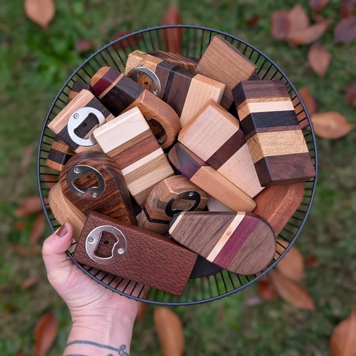 Handmade wooden bottle openers with metal openers in a black wire basket outdoors.