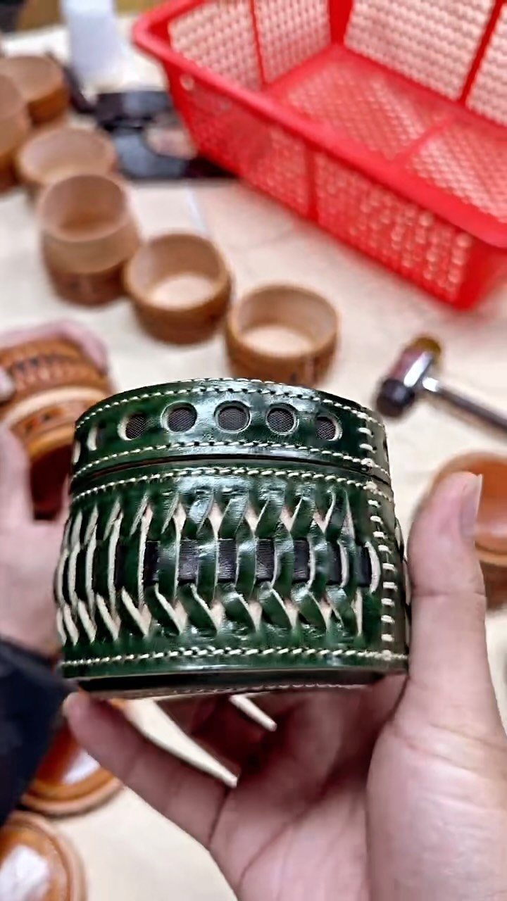 Green woven leather key basket held in hands with other leather crafts behind.