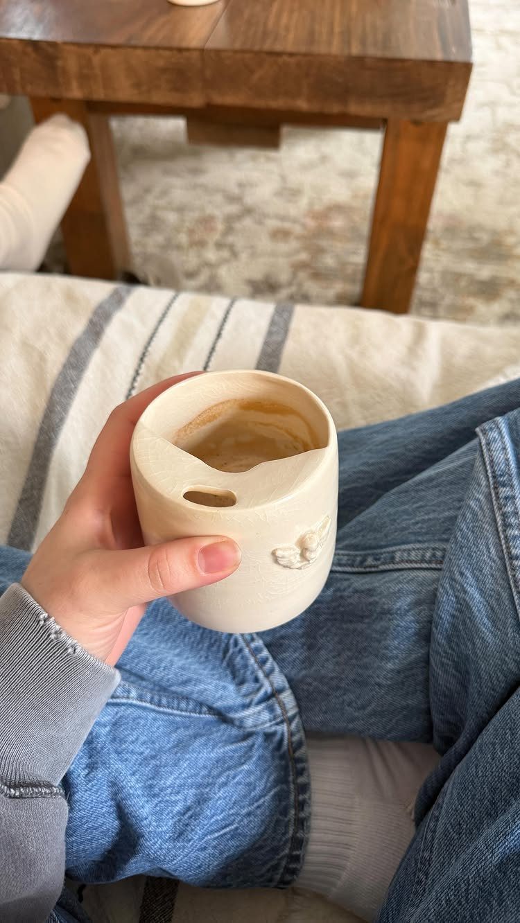 Handmade cream ceramic mug held by a person wearing blue jeans and grey sleeve.