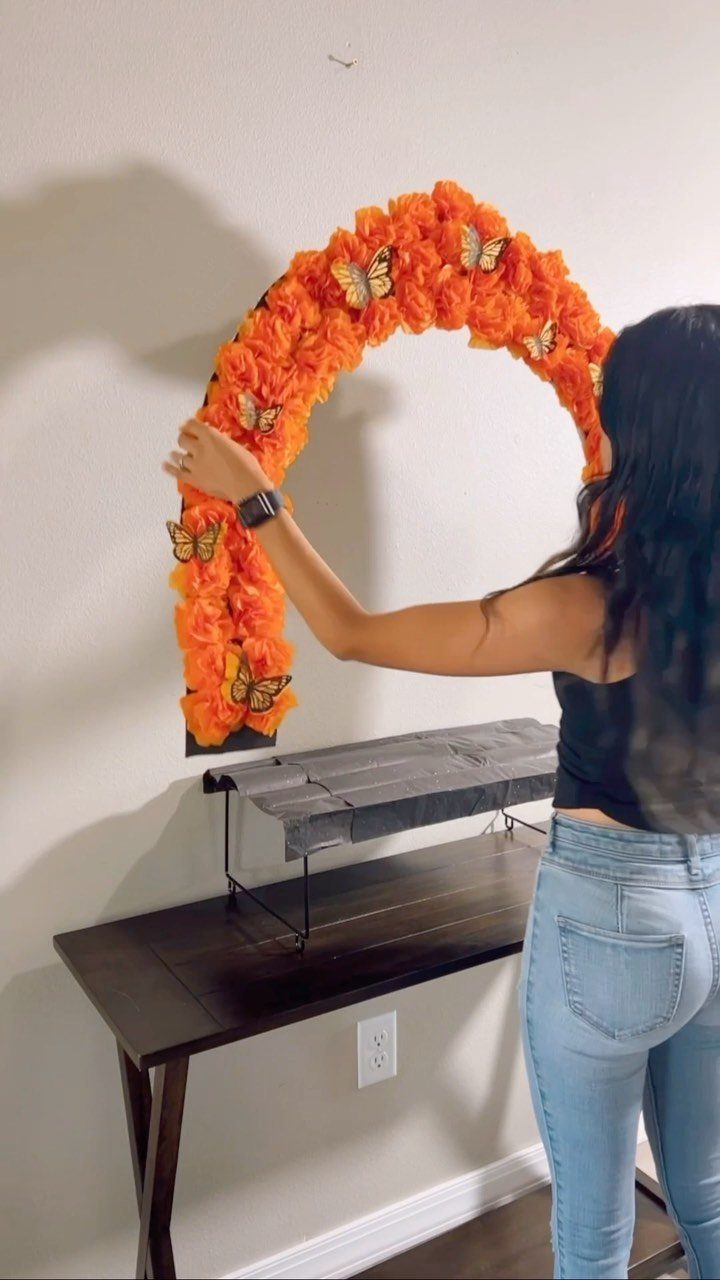 Woman adjusting an orange floral arch with butterflies on a white wall
