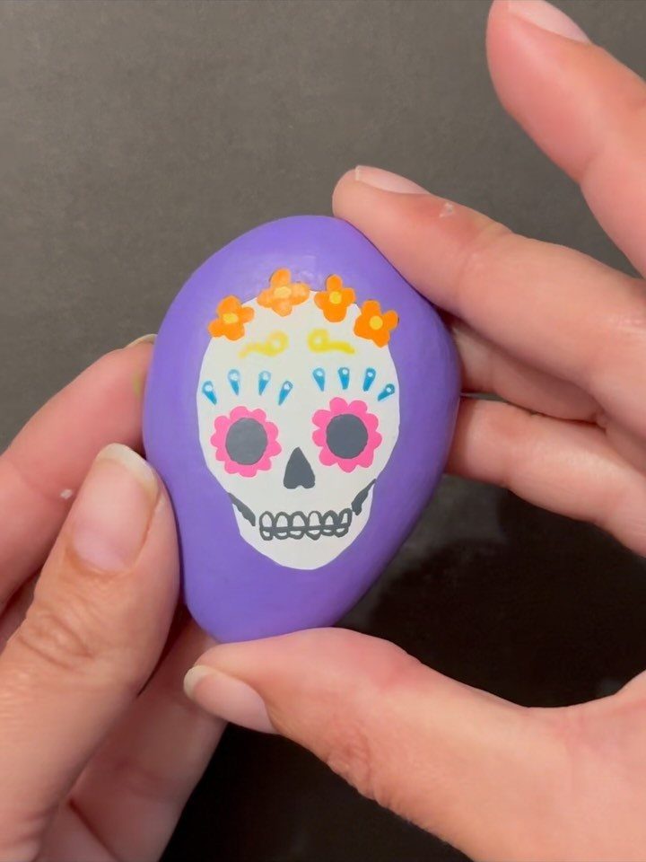 Purple painted rock with a colorful sugar skull design held by two hands.