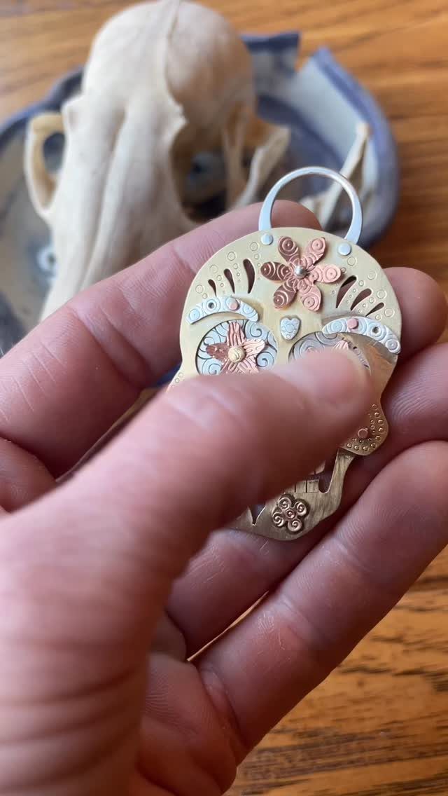 Gold metal sugar skull pendant with floral details held in a person's hand.