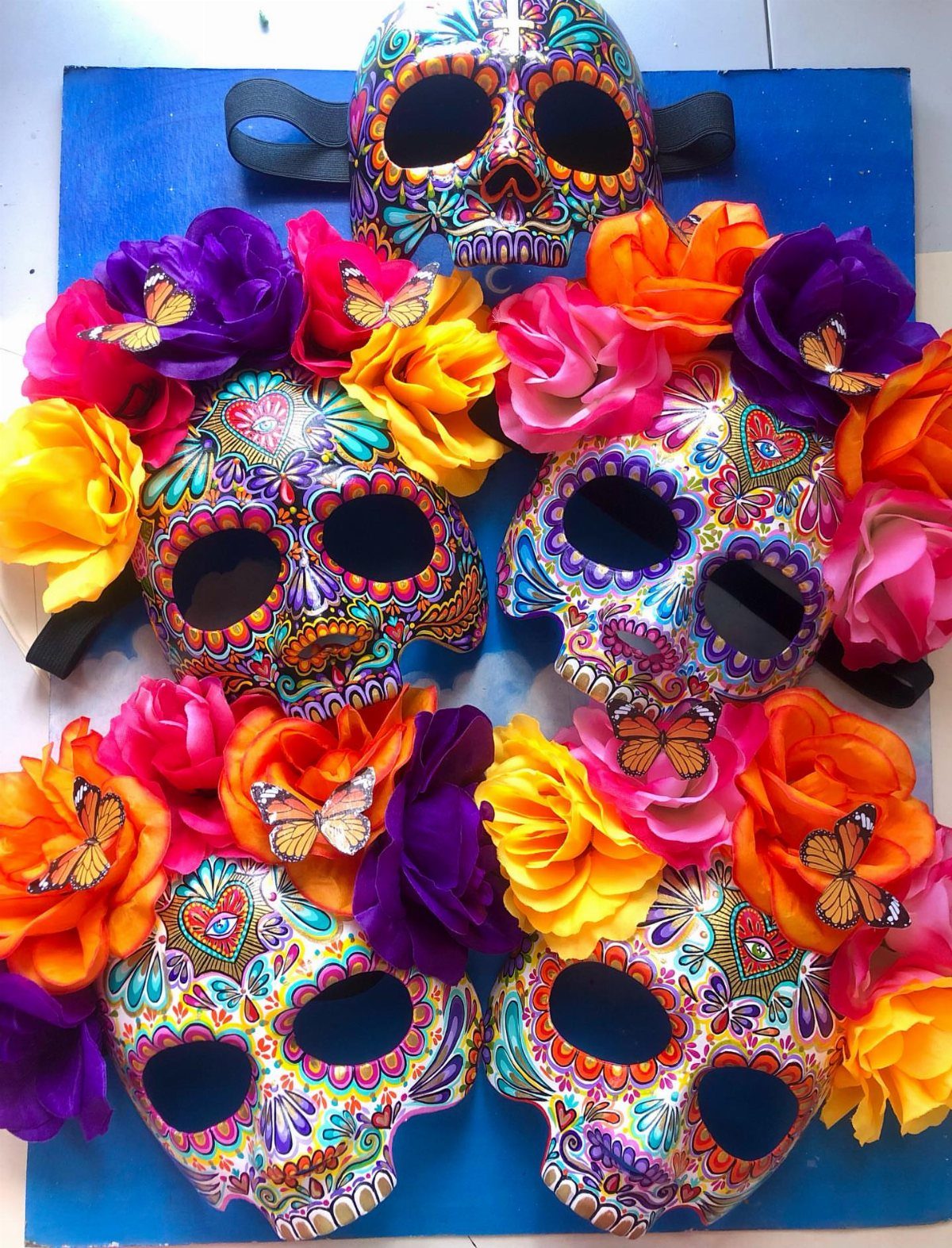 Four colorful sugar skull masks surrounded by bright artificial roses and butterflies.