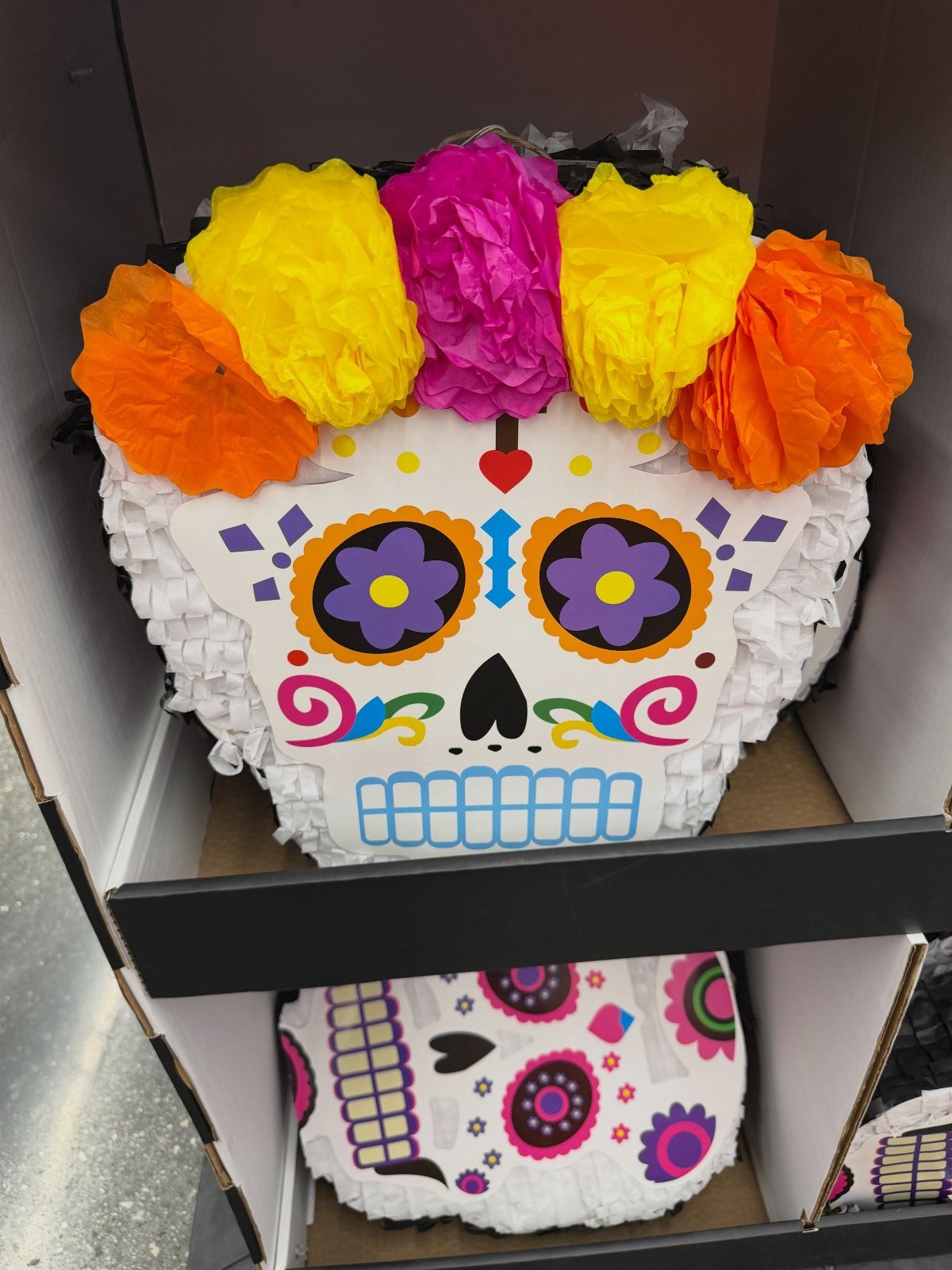 Sugar skull piñata with colorful tissue paper flowers in a retail box.