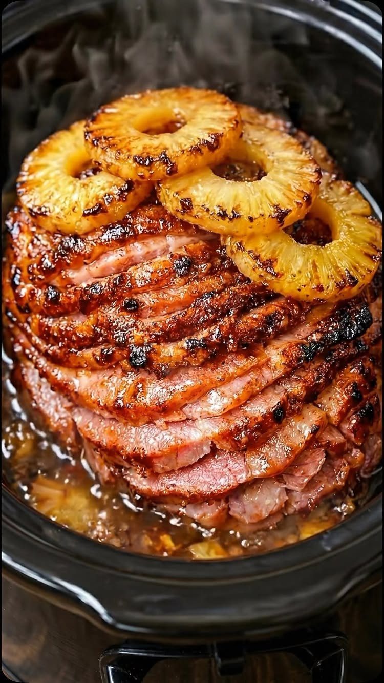 Glazed spiral-sliced ham topped with grilled pineapple rings in a black slow cooker.