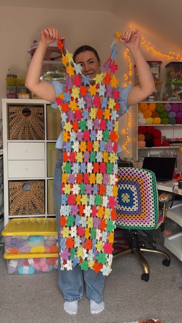 Woman holding a colorful handmade crochet dress in a room filled with yarn.