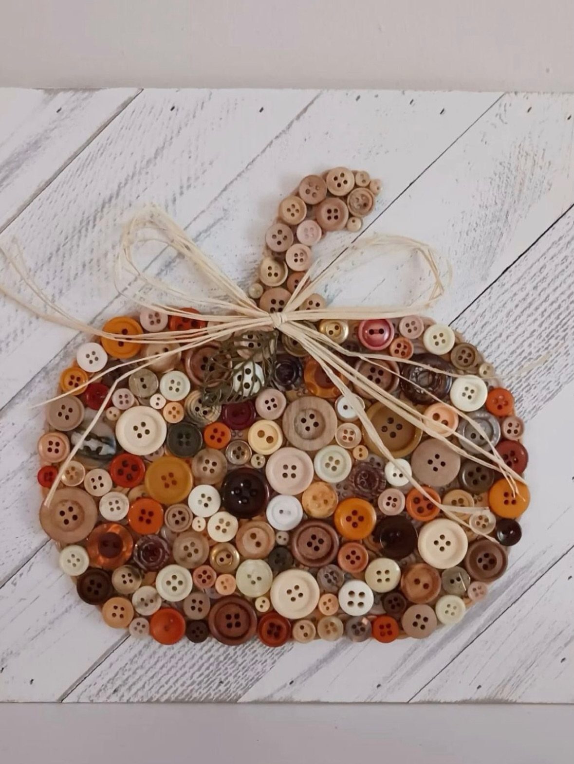 Button pumpkin art made of brown and orange buttons on white wood planks.