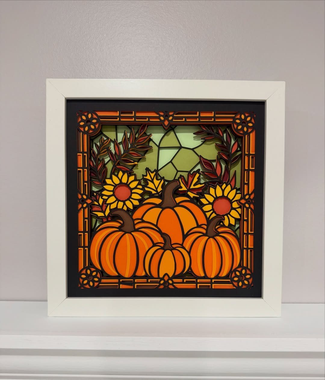 Layered paper art of pumpkins and sunflowers inside a white square shadow box frame.