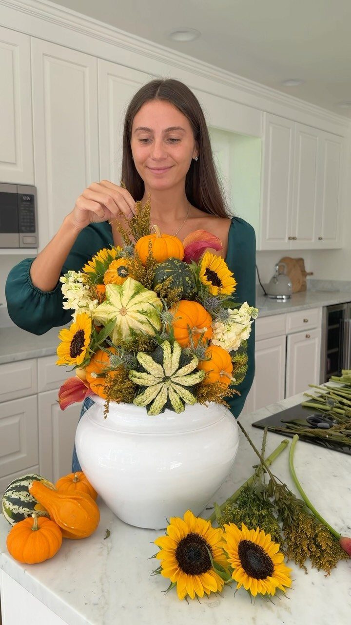 Woman arranging a fall centerpiece with sunflowers and pumpkins in a white vase.