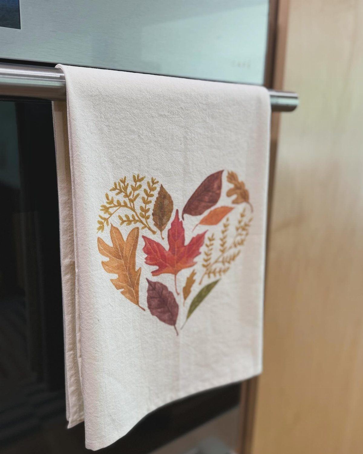 White tea towel with a heart made of autumn leaves hanging on oven handle