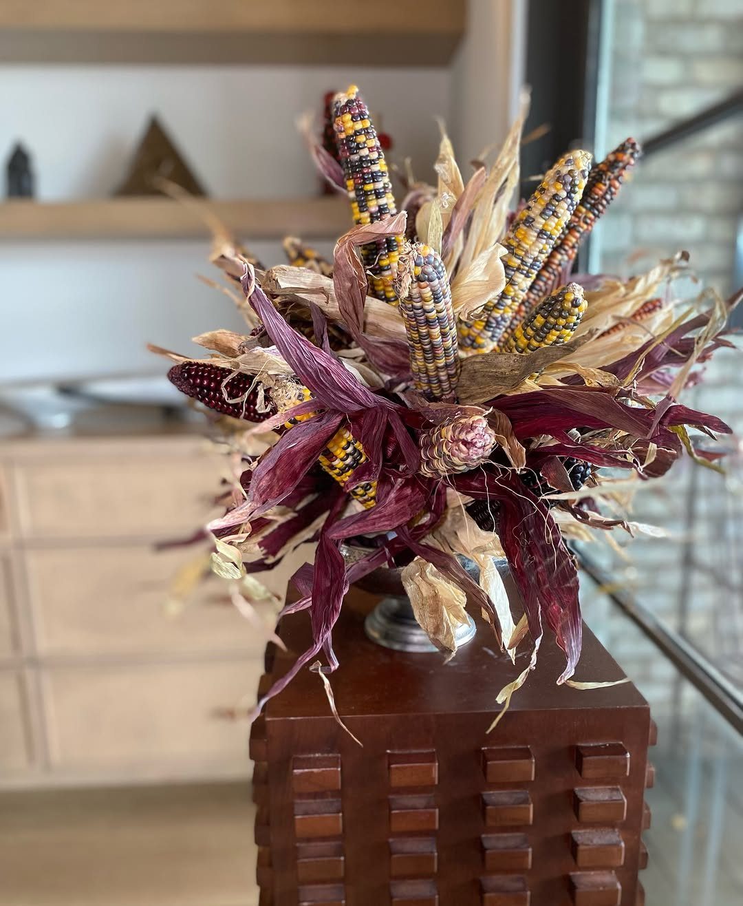 Ornamental maize corn arrangement in a silver vase on a wooden pedestal.