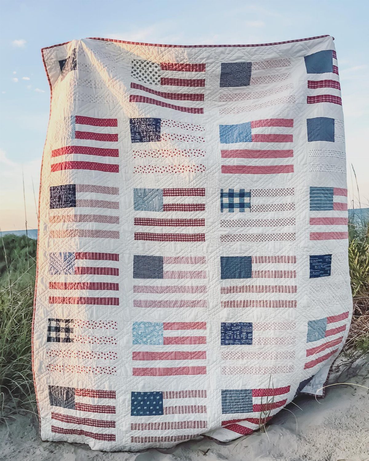 Quilted white blanket with various American flag patterns draped on a sandy beach.