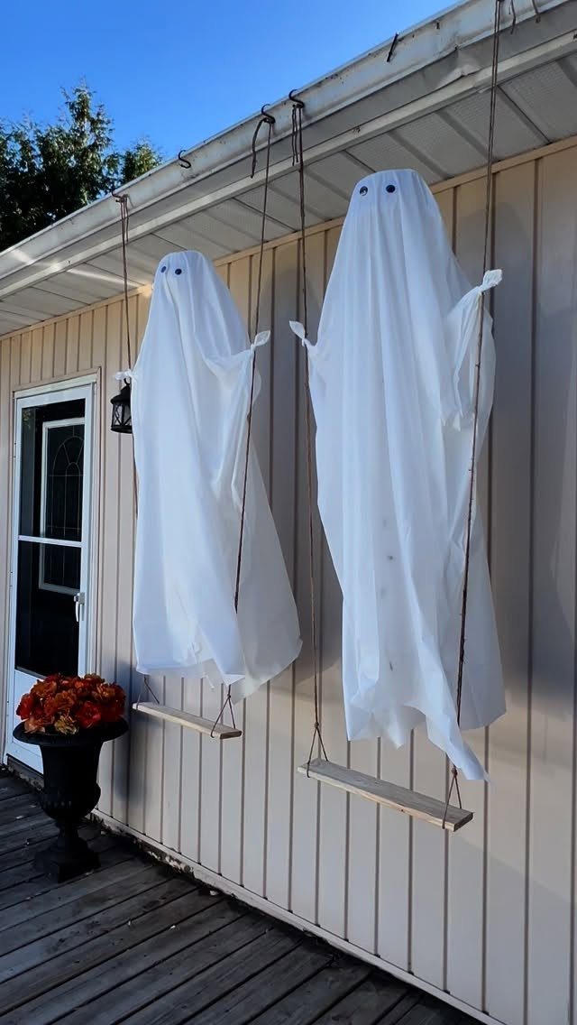 Two white sheet ghosts hanging on wooden swings on a house porch.