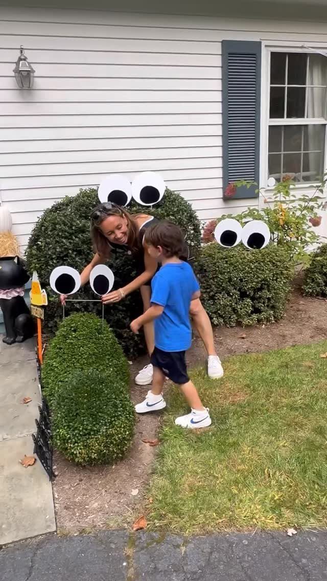 Woman and child placing large white eyes on green bushes for Halloween decor.