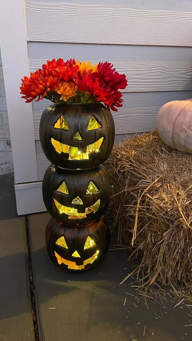 Stacked glowing black jack-o'-lanterns topped with red and orange flowers on a porch.