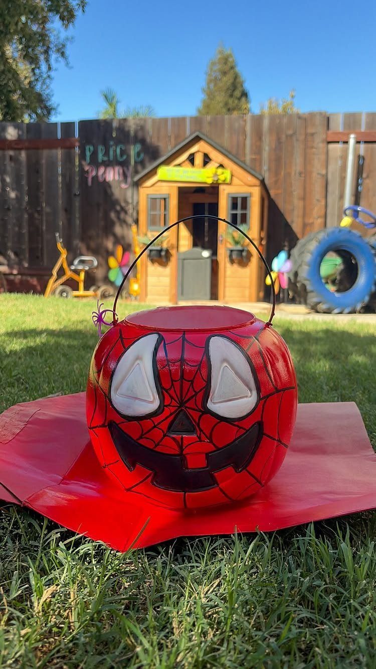 Spider-Man themed red plastic pumpkin bucket sitting on grass in a backyard