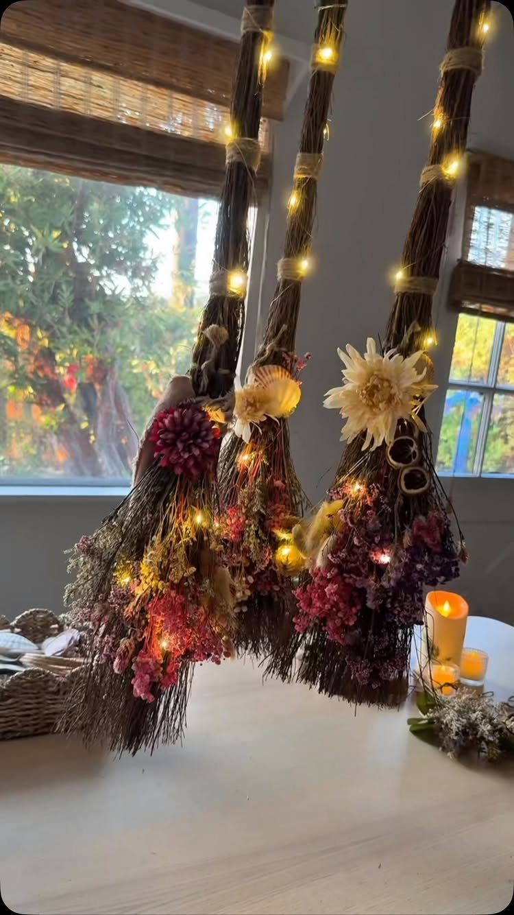 Three decorative witch's brooms with fairy lights and dried flowers on a table.
