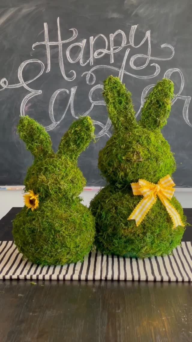 Two green moss bunny topiaries with yellow accents in front of an Easter chalkboard.