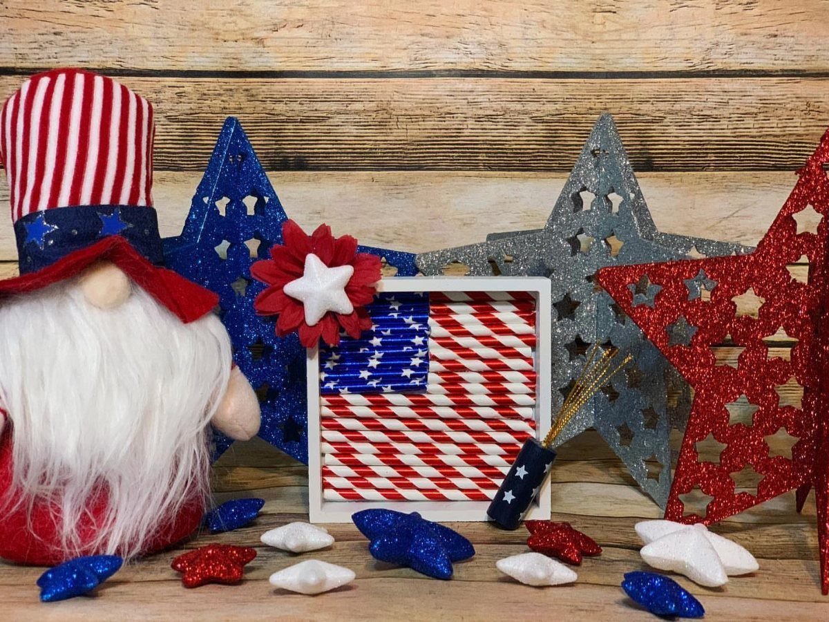 Patriotic gnome and glittery stars surrounding a straw American flag on wood.