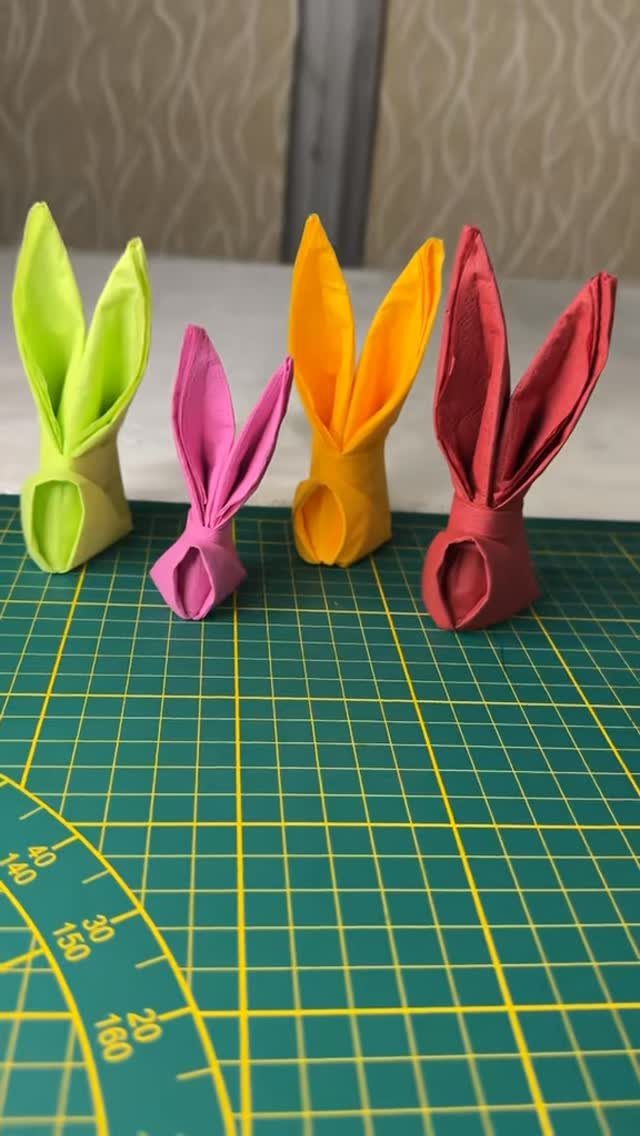 Four colorful cloth napkins folded into bunny shapes on a green cutting mat.