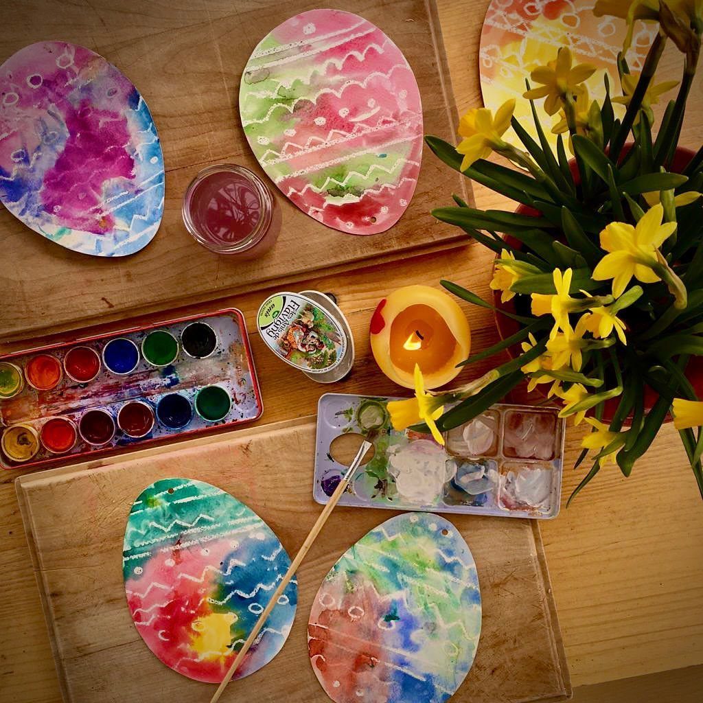 Colorful watercolor paper eggs with white resist patterns and painting supplies on wood.