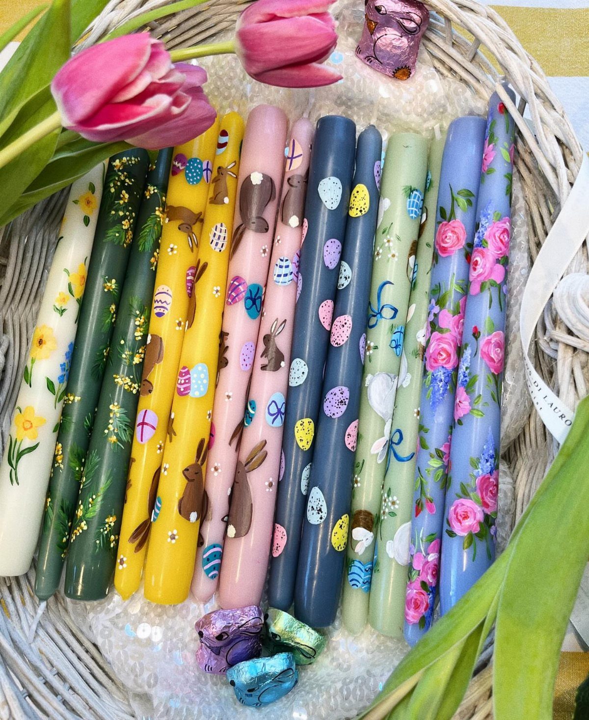 Painted taper candles with Easter motifs and flowers arranged in a white wicker basket.
