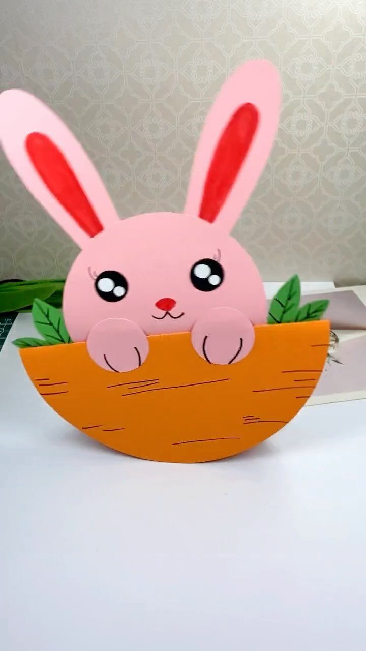 Pink paper bunny peeking out of an orange paper plate basket with green leaves.