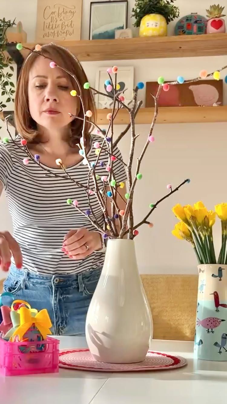 Woman decorating bare branches with colorful pastel pom-poms in a white vase.