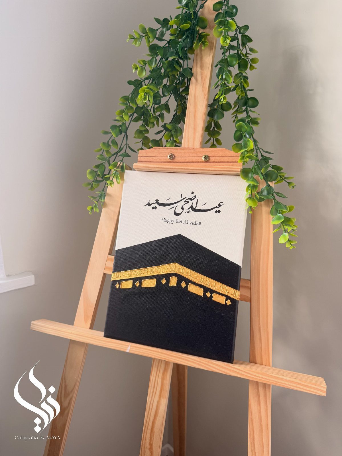 Arabic calligraphy painting of the Kaaba displayed on a wooden easel with greenery.