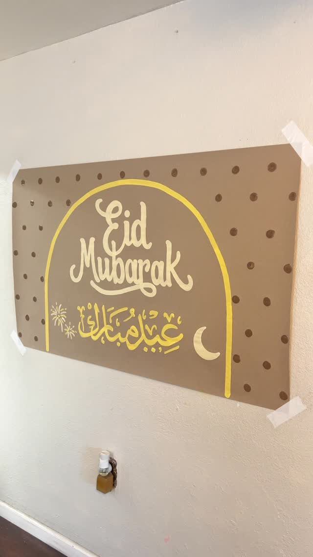 Brown polka dot banner with yellow Eid Mubarak calligraphy taped to a white wall